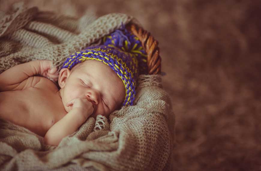תינוק שוכב ערום עם כובע ויד בפה על מצע סרוג